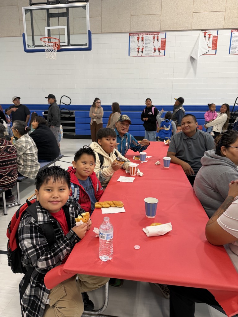 People seated at tables in a gymnasium. Tables have red covers, and some people hold food.