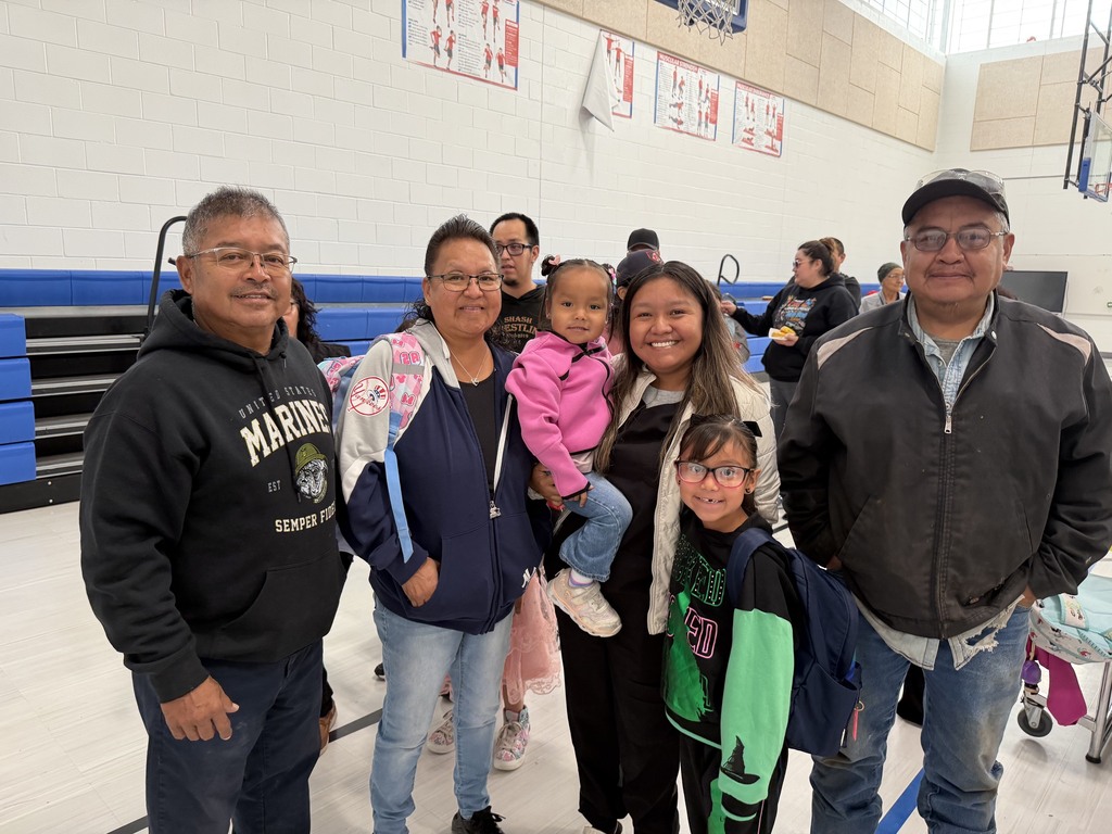 Five people in a gym with basketball equipment. A woman holds a child. Two adults are wearing glasses.