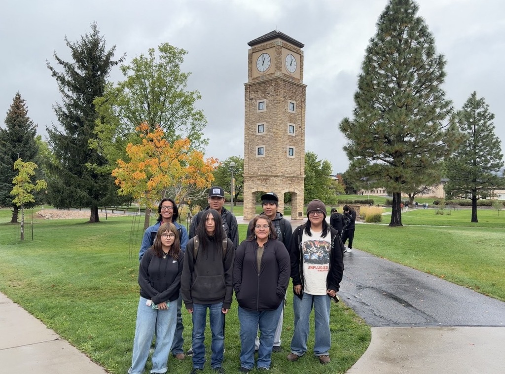 People stand on grass in front of a clock tower, with trees and a sidewalk behind them.