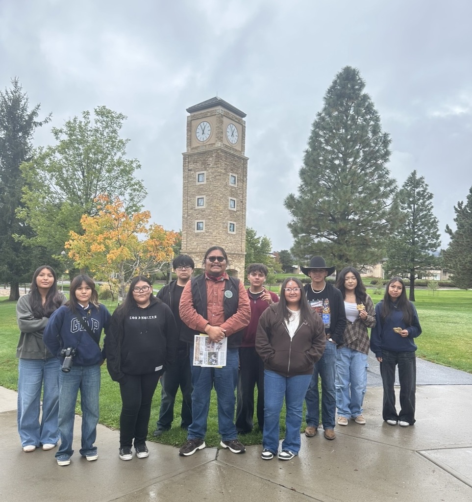 A group of people pose in front of a clock tower on a paved path. Trees and grass are in the background.