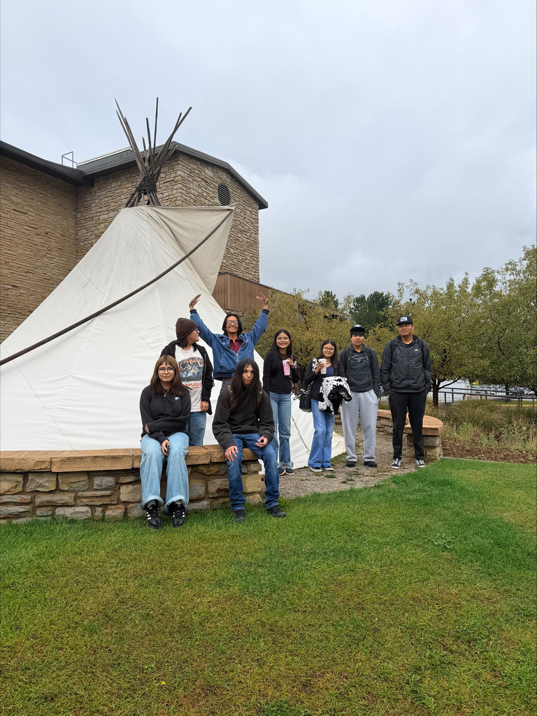 Group of people sitting and standing in front of a teepee with a stone wall and a brick building in the background.