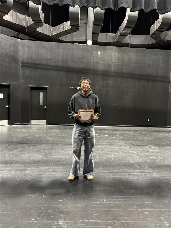 A person stands in a large, empty performance space holding a wooden award. Behind them, walls are black with two doors.