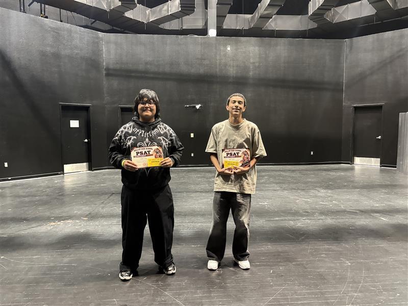 Two individuals stand in an empty room, each holding a box, with gray walls and a floor visible.