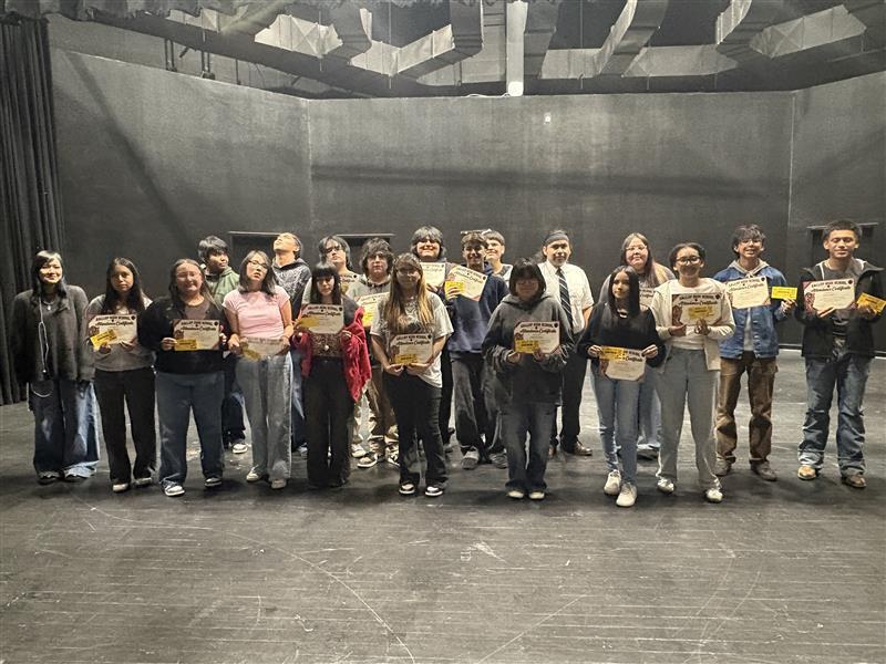 A group of people on a stage, holding certificates. Most wear casual clothing and sneakers. Behind them is a gray background with visible beams.
