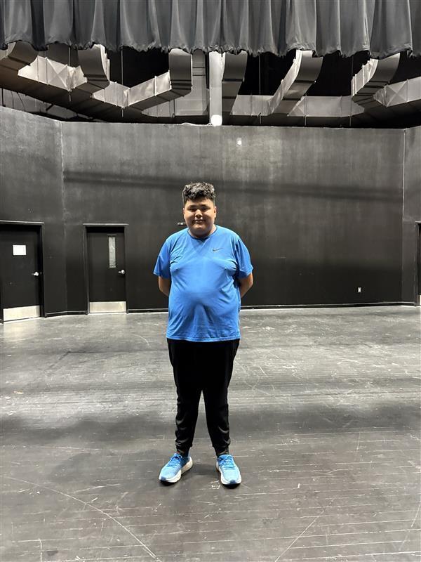 A person in a blue shirt and black pants stands in an empty auditorium with black walls and gray flooring.
