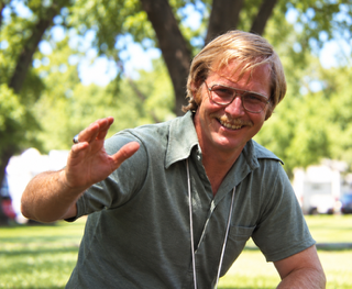 A person smiling, waving, and wearing a gray shirt. Behind them is a park with trees and a clear sky.