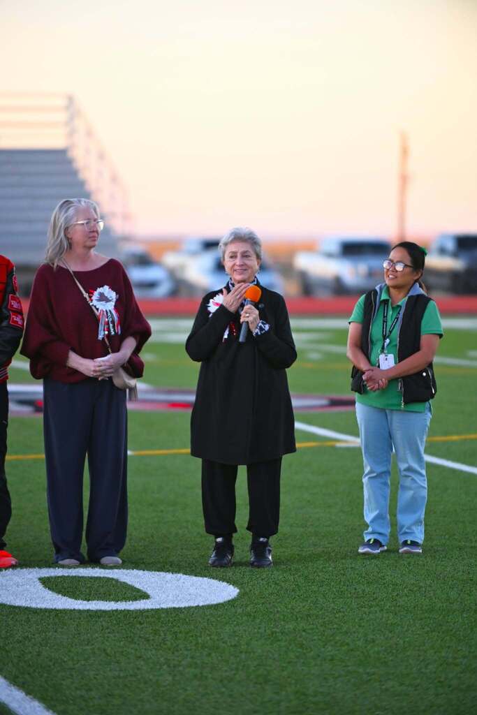 Three individuals stand on a football field with a sunset background. Each holds an object and appears to be waiting.