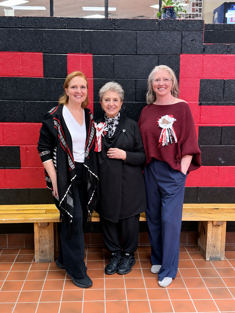 Three women in black, white, and maroon stand before a red and black wall. They wear jackets and pants.