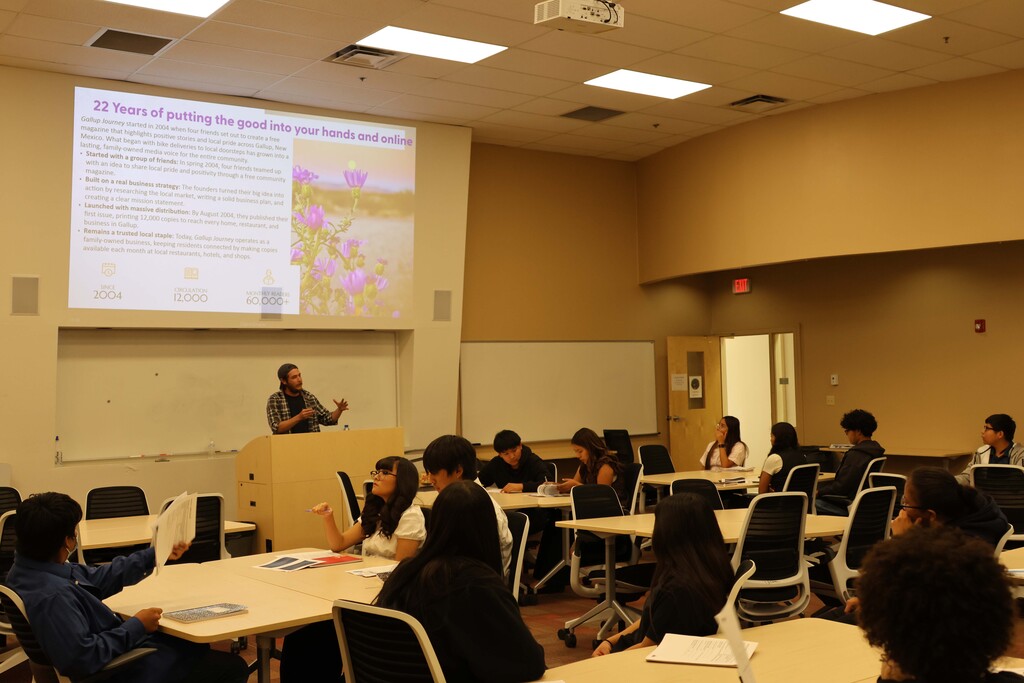 Room with projector screen showing presentation. Instructor at podium and students seated at tables.