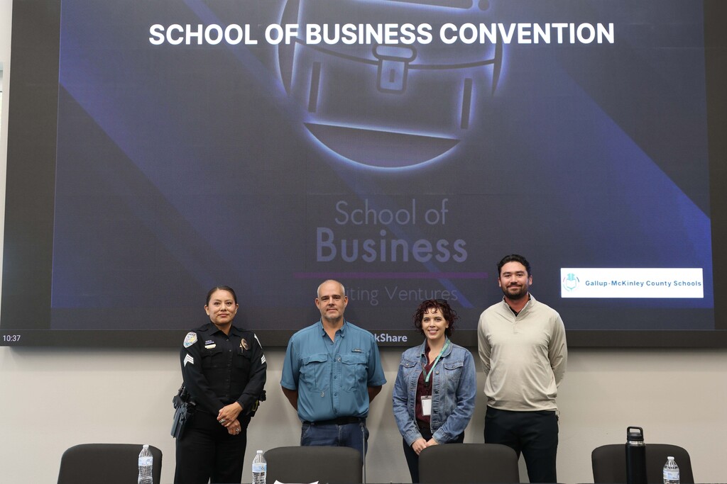 Four individuals stand in front of a screen displaying the words "School of Business Convention."