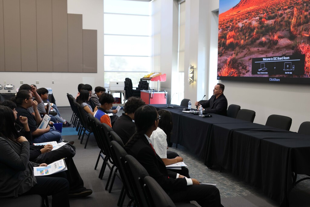 People seated in chairs in a classroom setting, listening to a man speak at a long table.