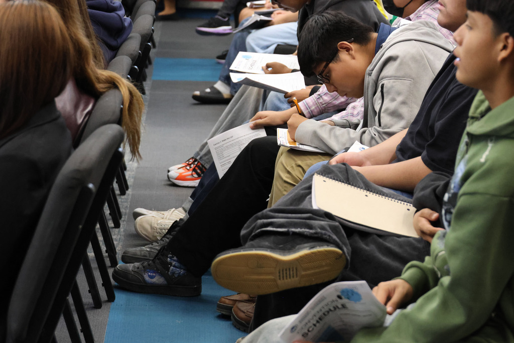 Several students in a classroom setting. Some are seated and writing on paper. Feet are visible.