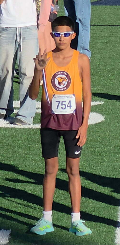 A boy wearing a sports jersey and shorts stands on a field with his hand raised. Number 754 is on his jersey.