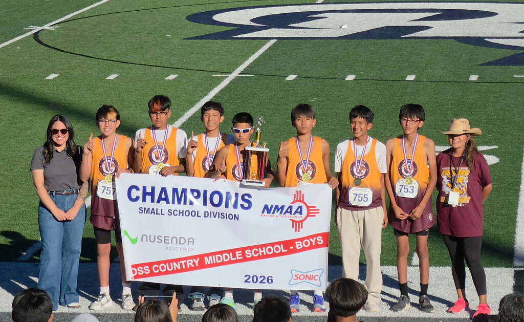 Students stand on a stage with a trophy and banner, celebrating a sports victory. They wear matching uniforms.