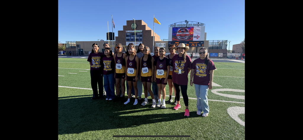 People in sports uniforms pose for a photo on a football field with a stadium in the background.