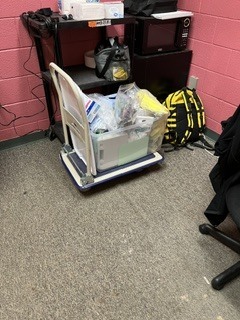 Cart with box of supplies on wheels, surrounded by bags and electronics. Pink wall in background.