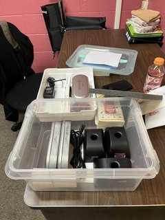 A plastic box on a table holds various items. A bottle, papers, and a black chair are nearby.