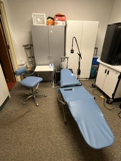 A room with blue reclining chair, a swivel chair, a desk, cabinets, a black monitor, a white box, and a black microphone on a stand.