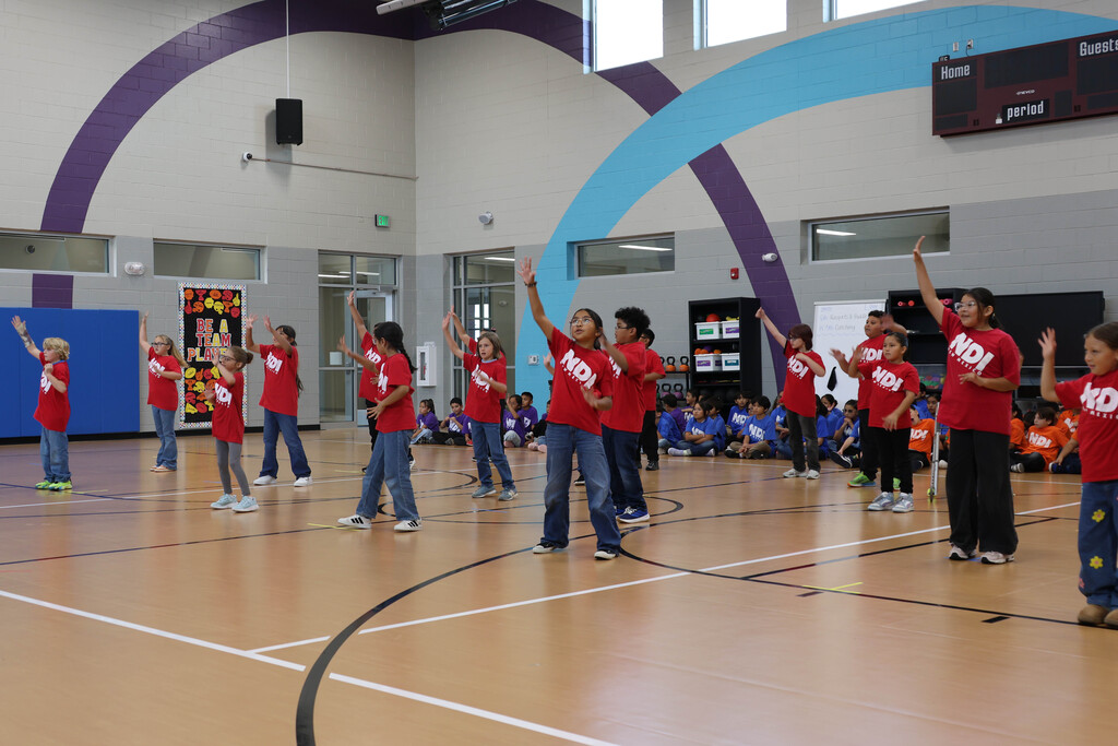 Several students wearing red shirts and blue jeans dance in an indoor gym. A scoreboard hangs from the ceiling.