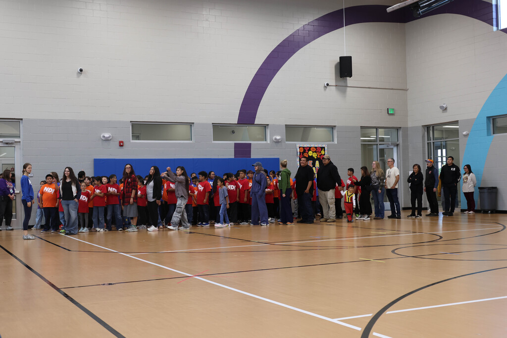 A group of people stand in a line inside a gym with a wooden floor and blue walls.