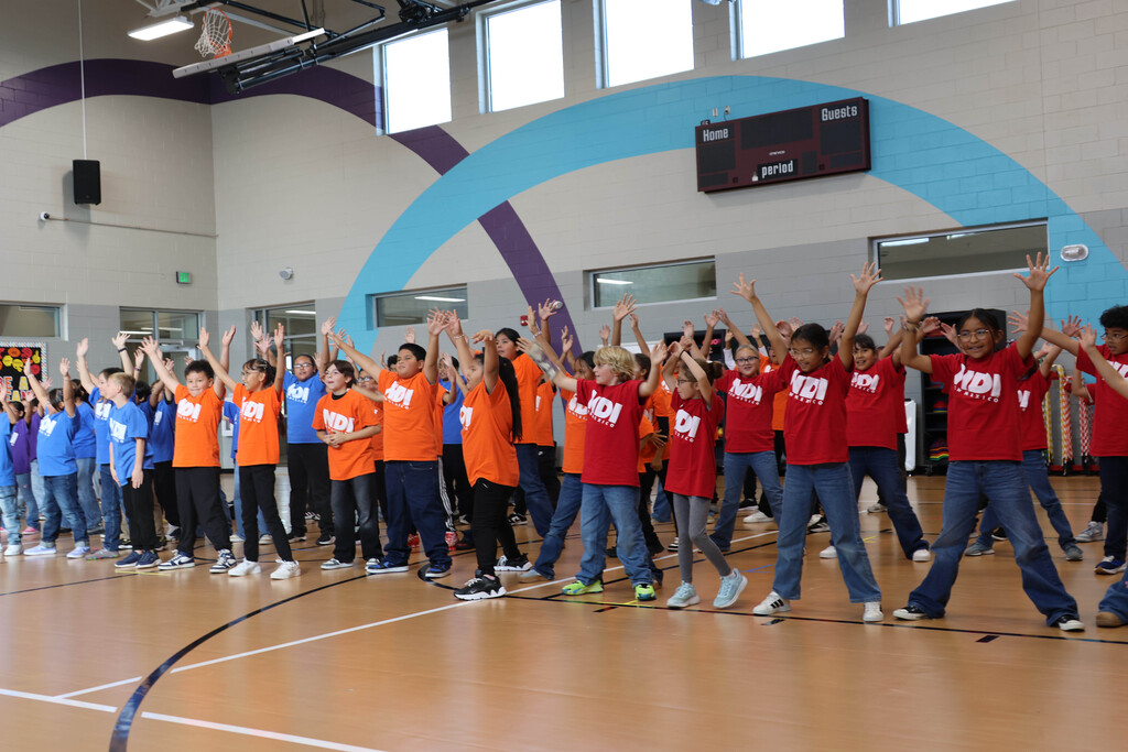 Students wearing orange shirts on a basketball court, standing with arms raised, facing a group of people.