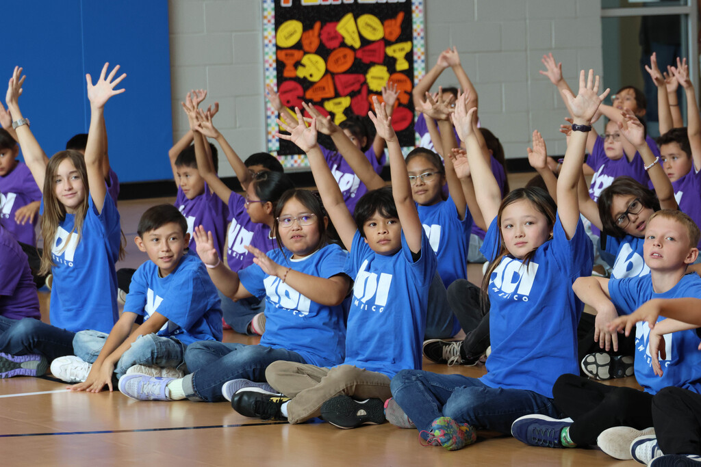 Group of children in blue shirts sitting on floor and raising their hands.