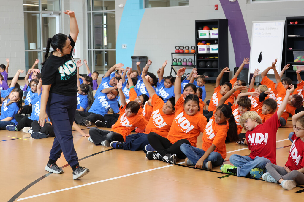 Woman leading a seated group of children in orange shirts; children raising arms, floor marked with lines.