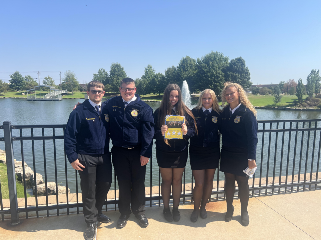 Five Gans FFA officers in blue FFA jackets stand together beside a pond and fountain at the COLT Conference.