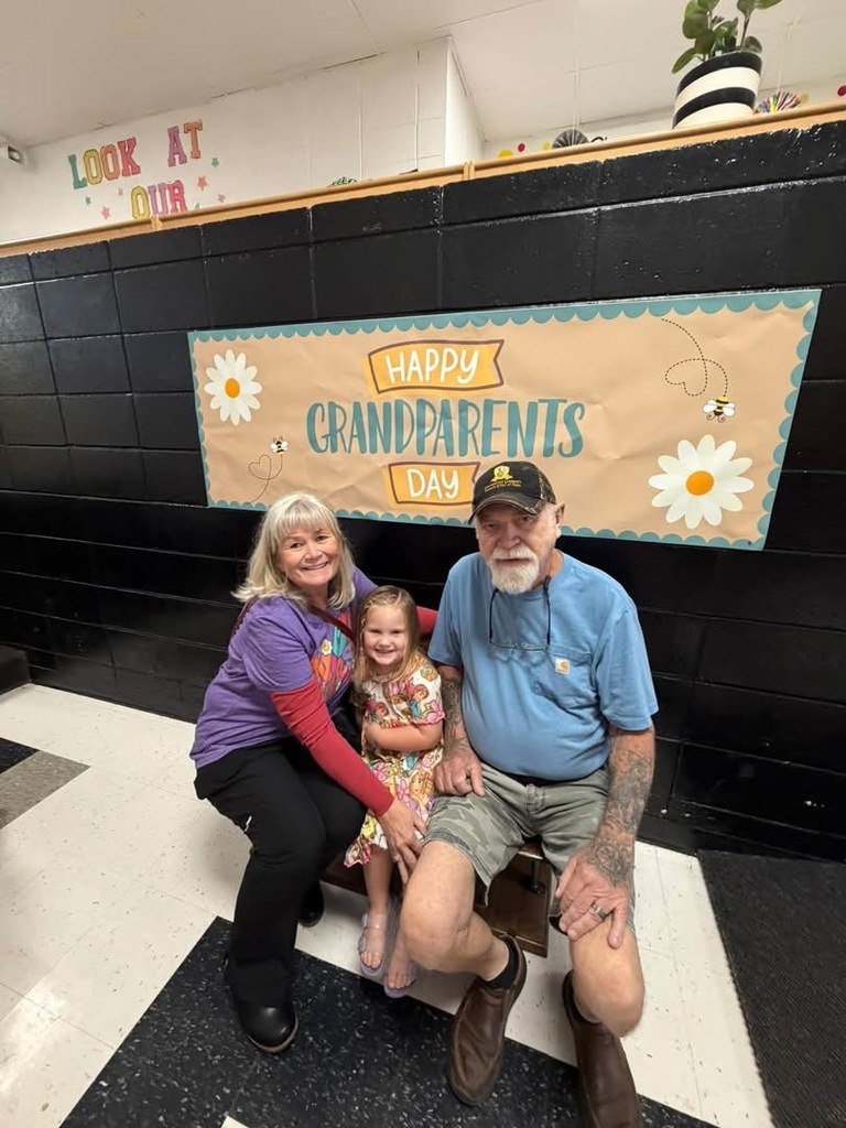A student sits with two grandparents beneath a Happy Grandparents Day banner.