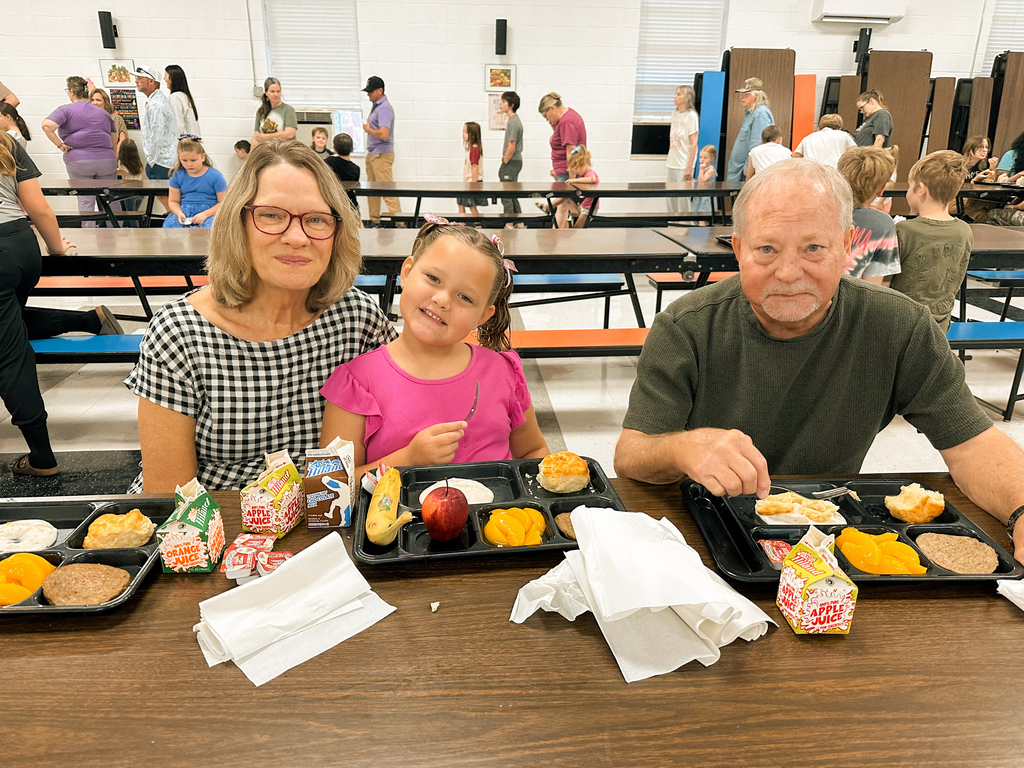A student smiles between two grandparents while enjoying breakfast in the school cafeteria.