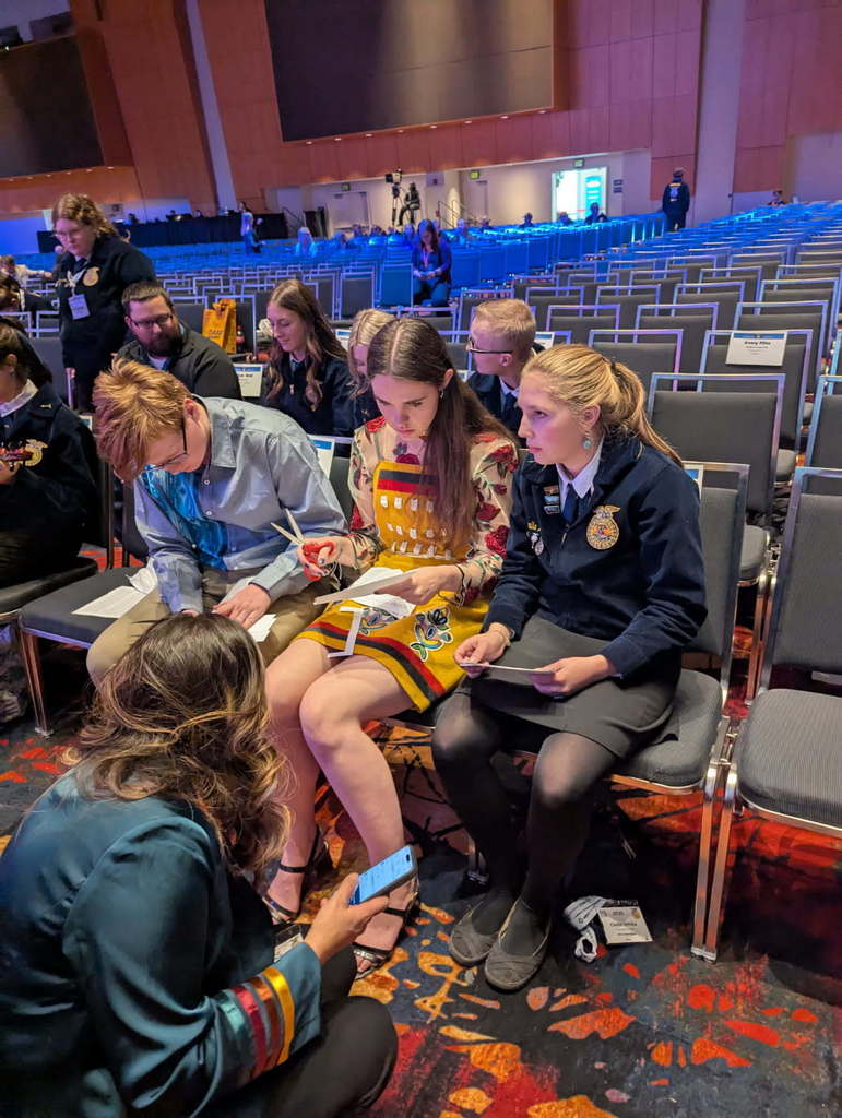 Three FFA members, sitting in chairs, prepare to present on stage along side their FFA advisor.