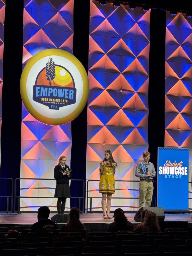Three FFA members stand on a stage at nationals, one wearing official FFA dress, one wearing a dress by a Native American fashion designer, and one wearing a modern ribbon shirt, giving a presentation.