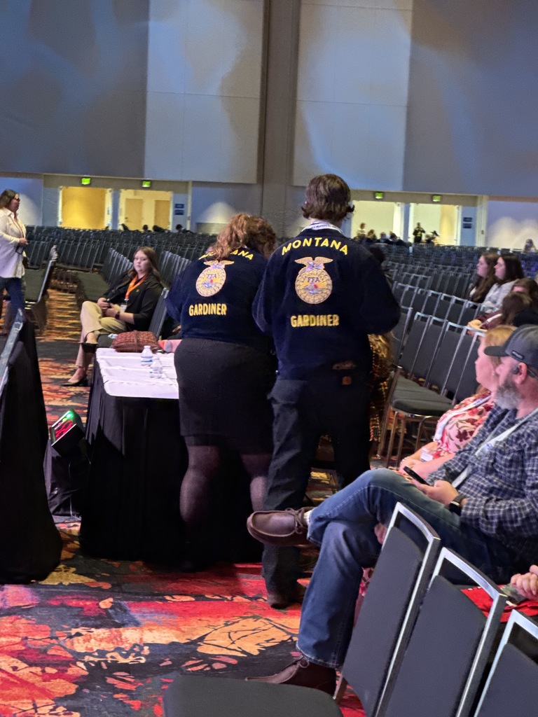 Two FFA members are seen handing items to judges seated at a table, with the perspective of looking at the back of the two FFA members, showing they are from Gardiner Montana.