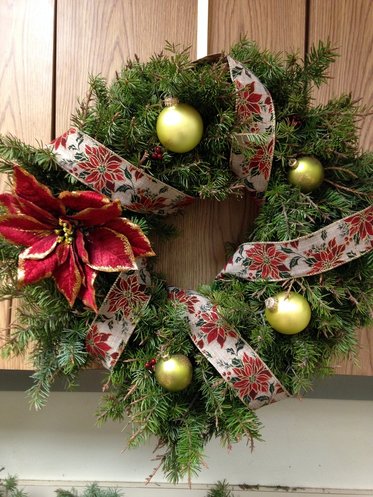Christmas wreath on display with a red poinsettia, ribbon , and gold bulbs. 