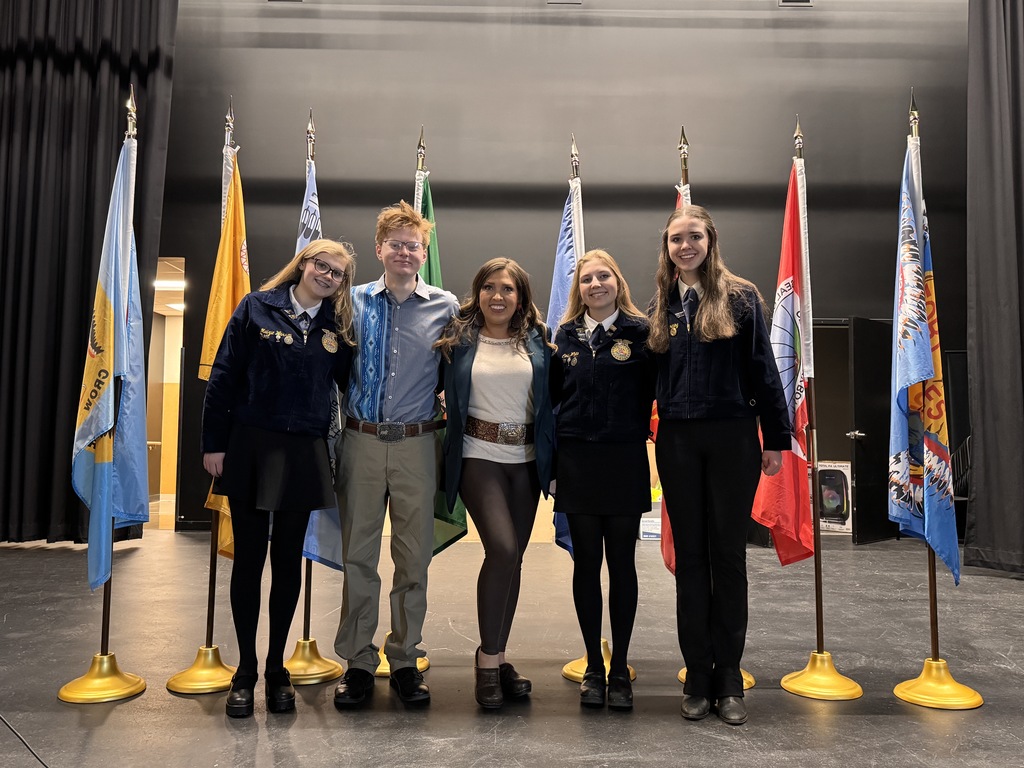 Four FFA members stand with FFA advisor Mrs. Hoppe on a stage with the tribal flags of Montana displayed on stands behind them.