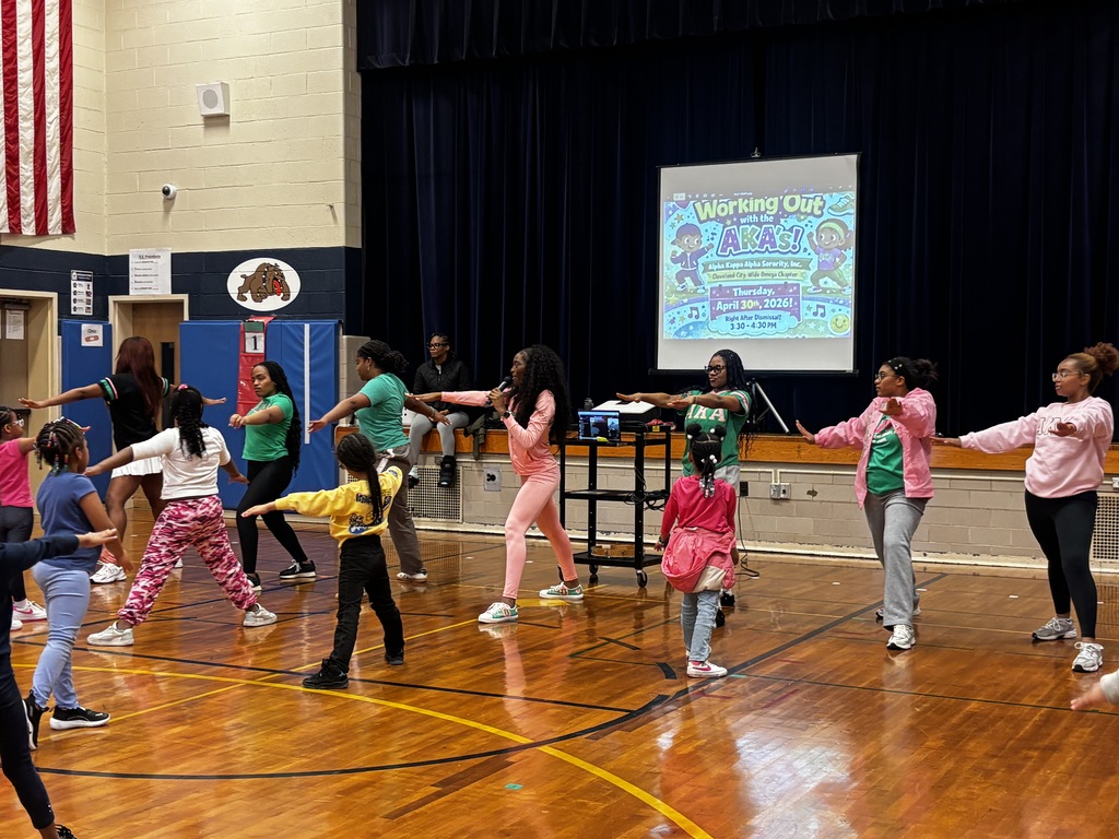 A group of women in pink and green athletic wear lead a fitness session on a wooden gymnasium floor. In the center, a woman in a pink tracksuit speaks into a microphone while demonstrating a side-stretch pose. To her right, several women in green "AKA" shirts and pink jackets mirror the movement. A group of elementary-aged children follow along in the foreground. A projector screen in the background displays a colorful graphic that reads "Working Out with the AKAs!" and the date "April 30th, 2026."