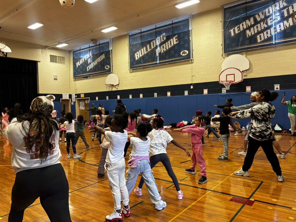 A side-view of the gymnasium showing rows of children practicing a coordinated arm-reaching exercise. The children are focused, following the lead of instructors scattered throughout the room. The gym walls are decorated with blue padding and motivational banners that read "Bulldog Pride" and "Teamwork Makes the Dream Work." The lighting is bright, reflecting off the polished wood floors.