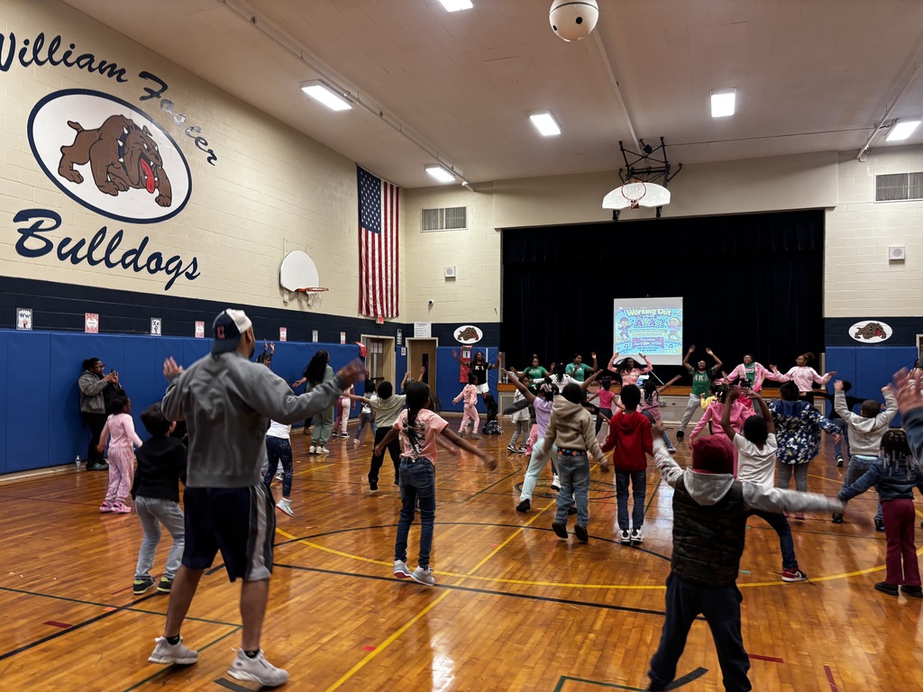 A wide-angle view from the back of the gym during the "Working Out with the AKAs" event. A large group of children and several adults are seen from behind, performing jumping jacks in unison. The gym walls feature a large "William Foster Bulldogs" logo and a prominent American flag. At the far end of the court, the instructors are on a stage area in front of the event's digital flyer, leading the high-energy session.