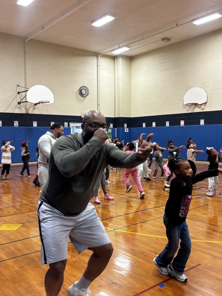 A close-up action shot in a brightly lit gym showing a tall man with glasses and a grey hoodie leading a workout. He is in a wide stance, throwing a controlled punch toward the camera with a focused expression. To his right, a young girl in a black shirt mimics his boxing stance with a big smile. In the background, dozens of other children and adults are spread across the basketball court, all participating in the same exercise.