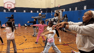 A looping clip of a large group of students and instructors in a gymnasium performing a synchronized yoga stretch. They hold a deep side lunge with arms extended horizontally, slowly lowering their arms in unison as they practice mindful breathing.