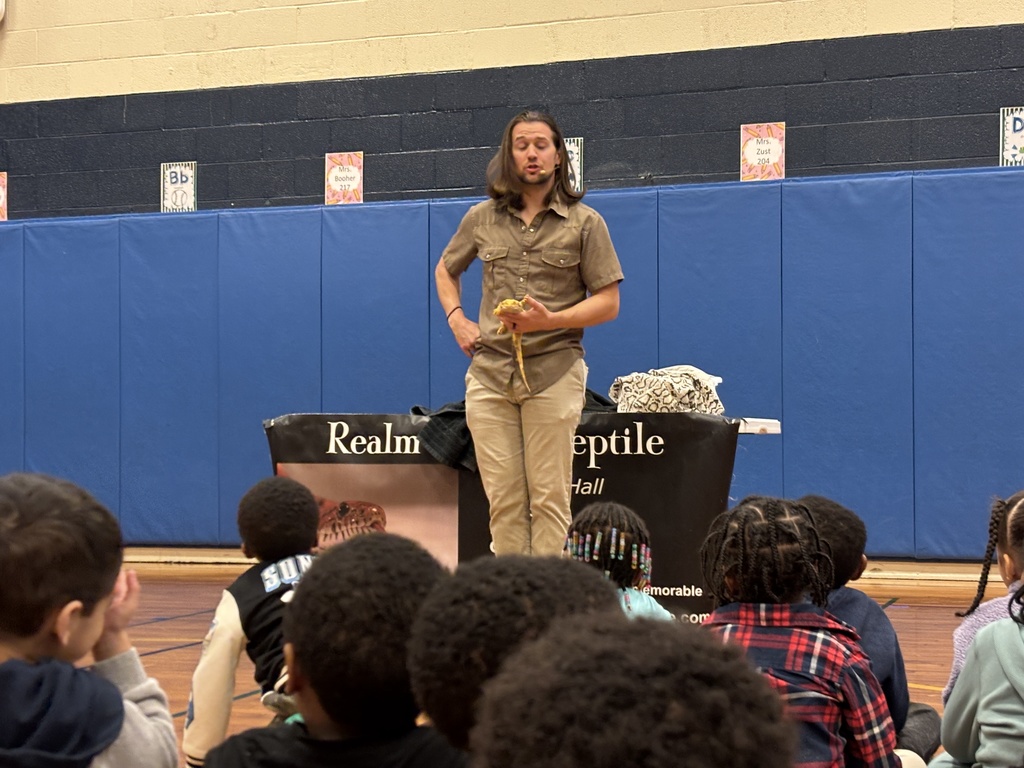 A wide shot in a school gymnasium where a male presenter with long brown hair, wearing a tan button-down shirt and khakis, stands before a seated audience of young children. He is holding a small, yellow-orange lizard (bearded dragon) in his hands. Behind him is a blue padded wall and a table draped with a black banner that reads "Realm of the Reptile