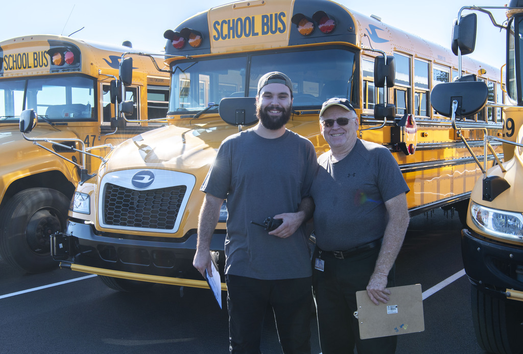 2 members of the transportation team helping coordinate the bus move, smiling in front of buses
