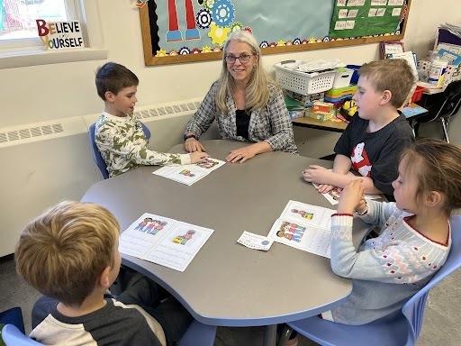Image of Mrs. Maust, seated at a table with four students, assisting with their learning.