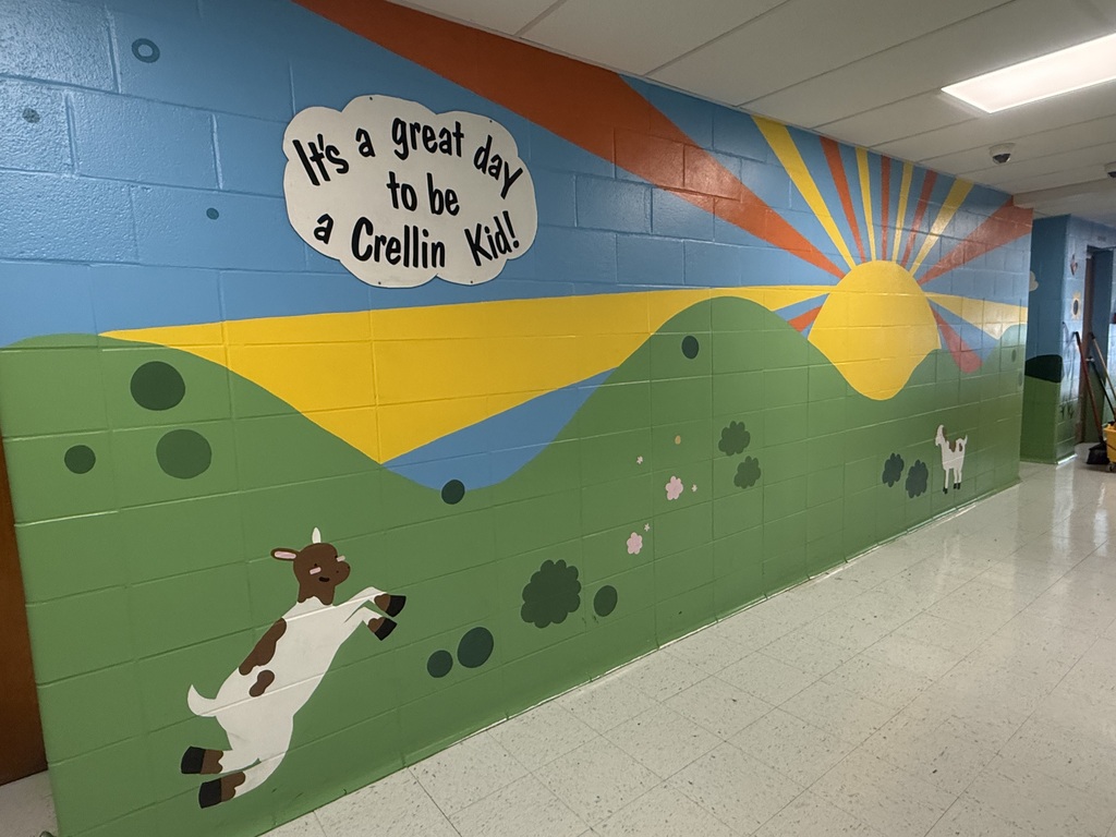 A mural is painted on a wall representing outdoor aspects of Crellin Elementary School.  The mural features a goat leaping in a meadow, with the sun glowing from the horizon.