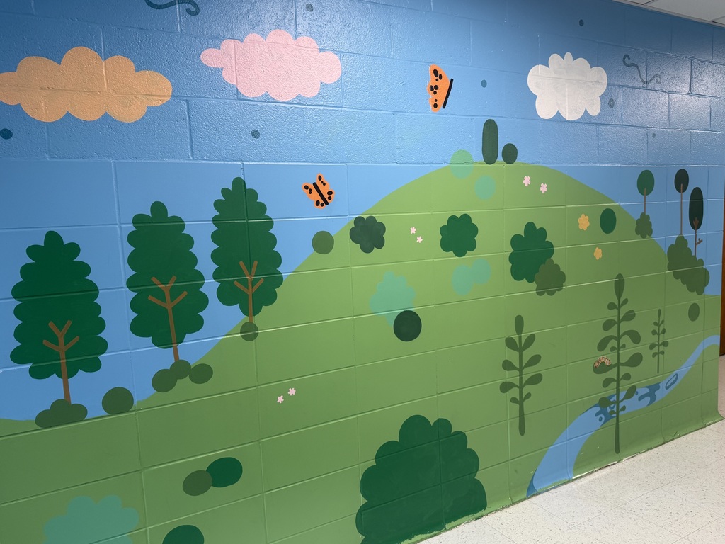 A mural is painted on a wall representing outdoor aspects of Crellin Elementary School.  The mural features a stream at the base of a hill, with trees and butterflies.