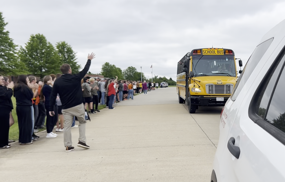 Many people line a street; a school bus is moving past them. A person waves to the bus.