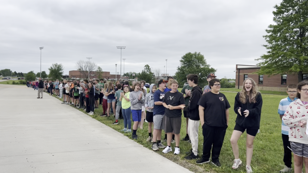 A line of students stands on grass, facing a building with a brick wall and trees.