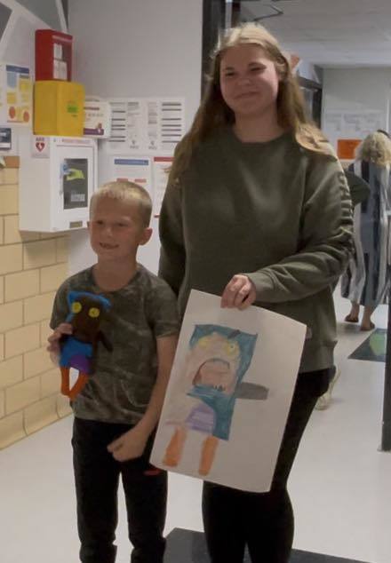 Woman in green sweater and child in camouflage shirt standing in a hallway with a drawing and stuffed toy.