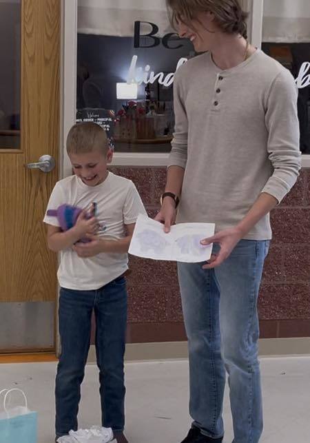 Boy in white shirt holds gift; adult in tan shirt presents paper. Both stand in front of door, with store window behind.
