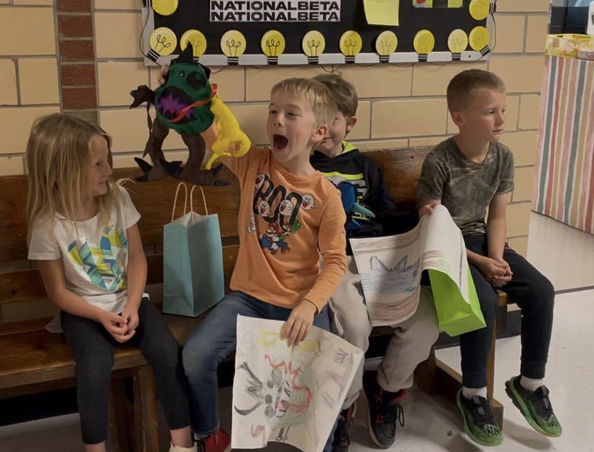 Five kids on a bench, one holding a yellow object and shouting, others holding papers. A colorful banner is in the background.